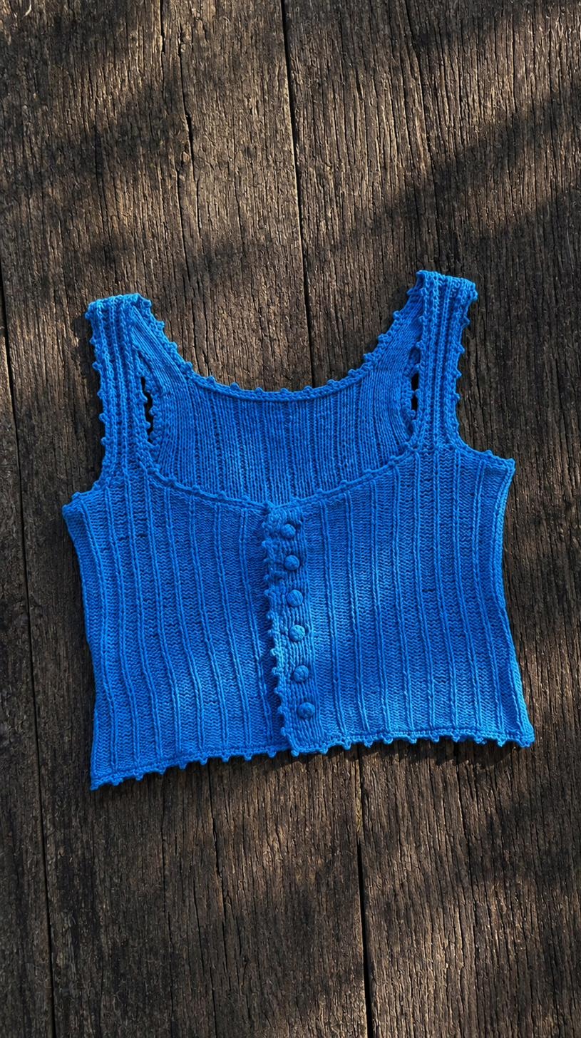 Blue ribbed hand-knitted tank top with covered buttons, laid flat on dark wood